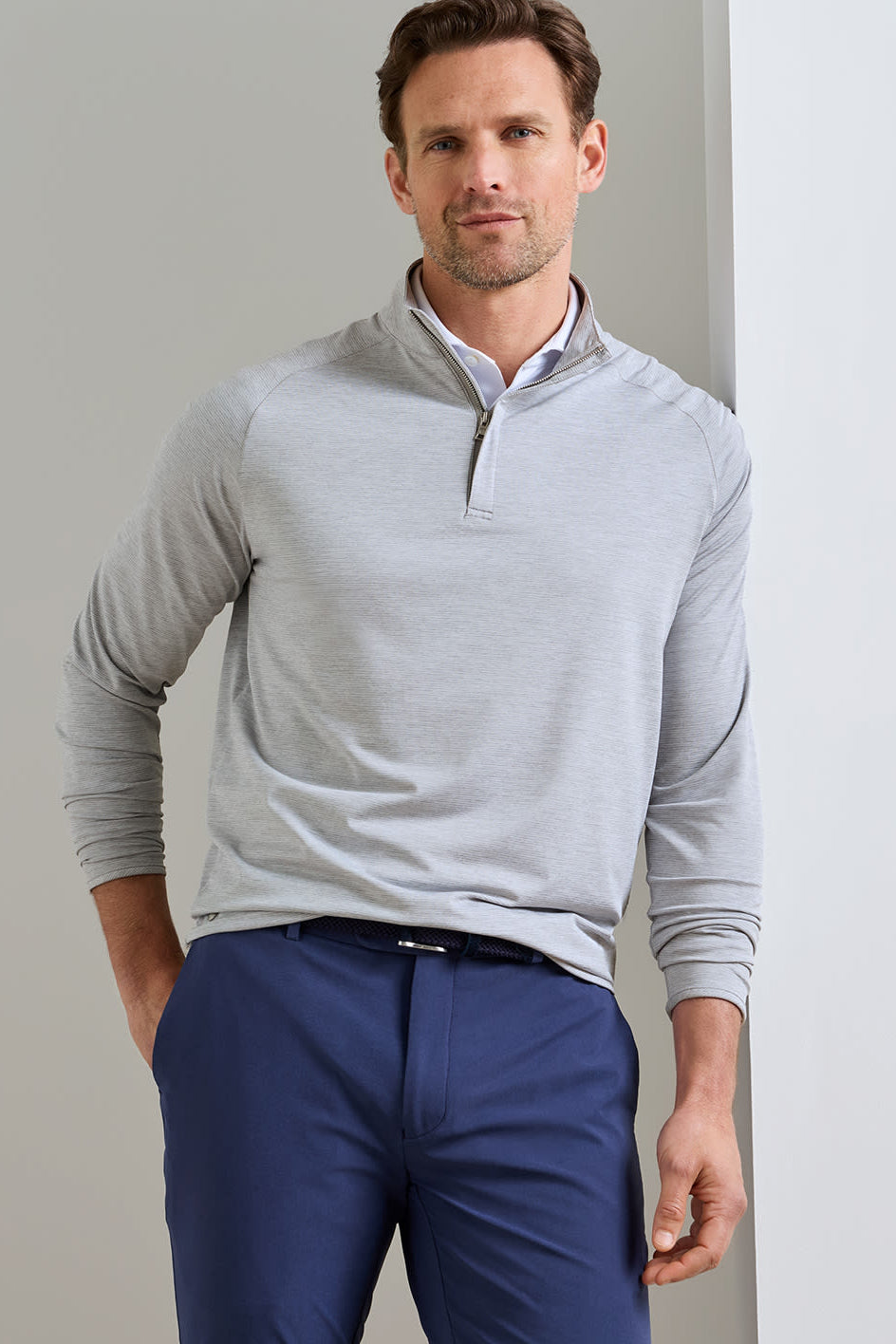 A man wearing a light gray quarter-zip long-sleeve pullover over a white collared shirt and navy blue trousers, standing against a neutral light gray wall.