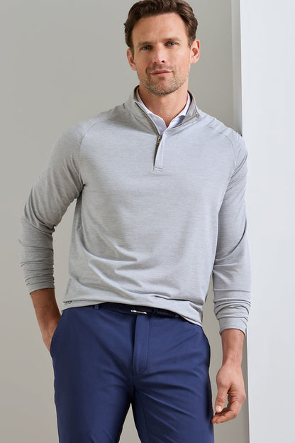 A man wearing a light gray quarter-zip long-sleeve pullover over a white collared shirt and navy blue trousers, standing against a neutral light gray wall.