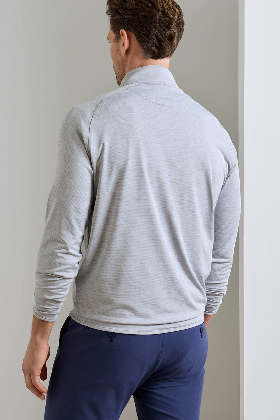 A man viewed from behind wearing a light gray heathered quarter-zip mock neck pullover and navy blue trousers, standing against a neutral light gray wall.