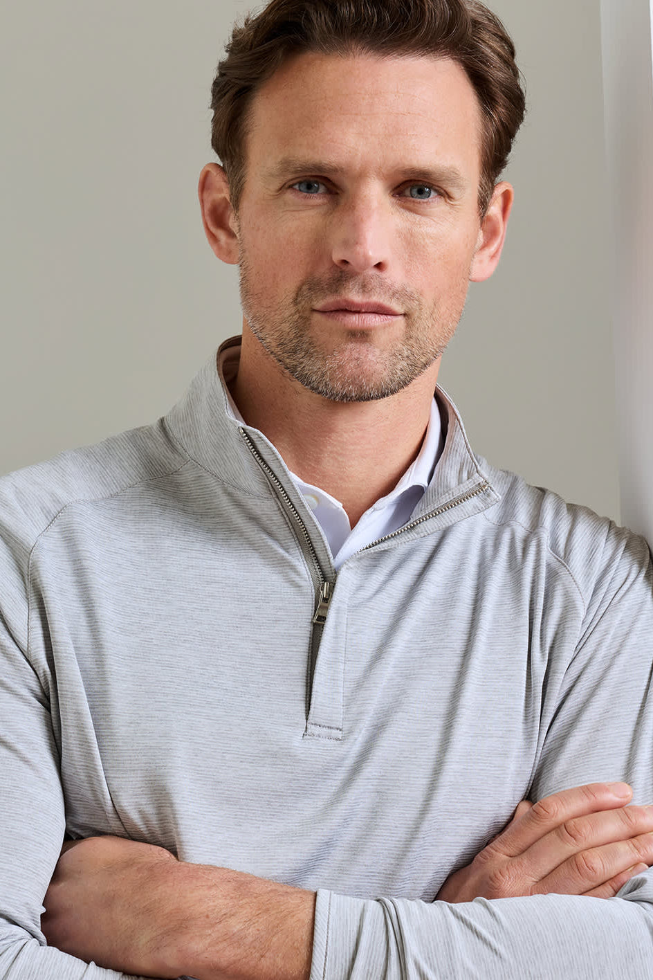 A man wearing a light gray quarter-zip long-sleeve pullover over a white collared shirt, standing with arms crossed against a neutral light gray background.