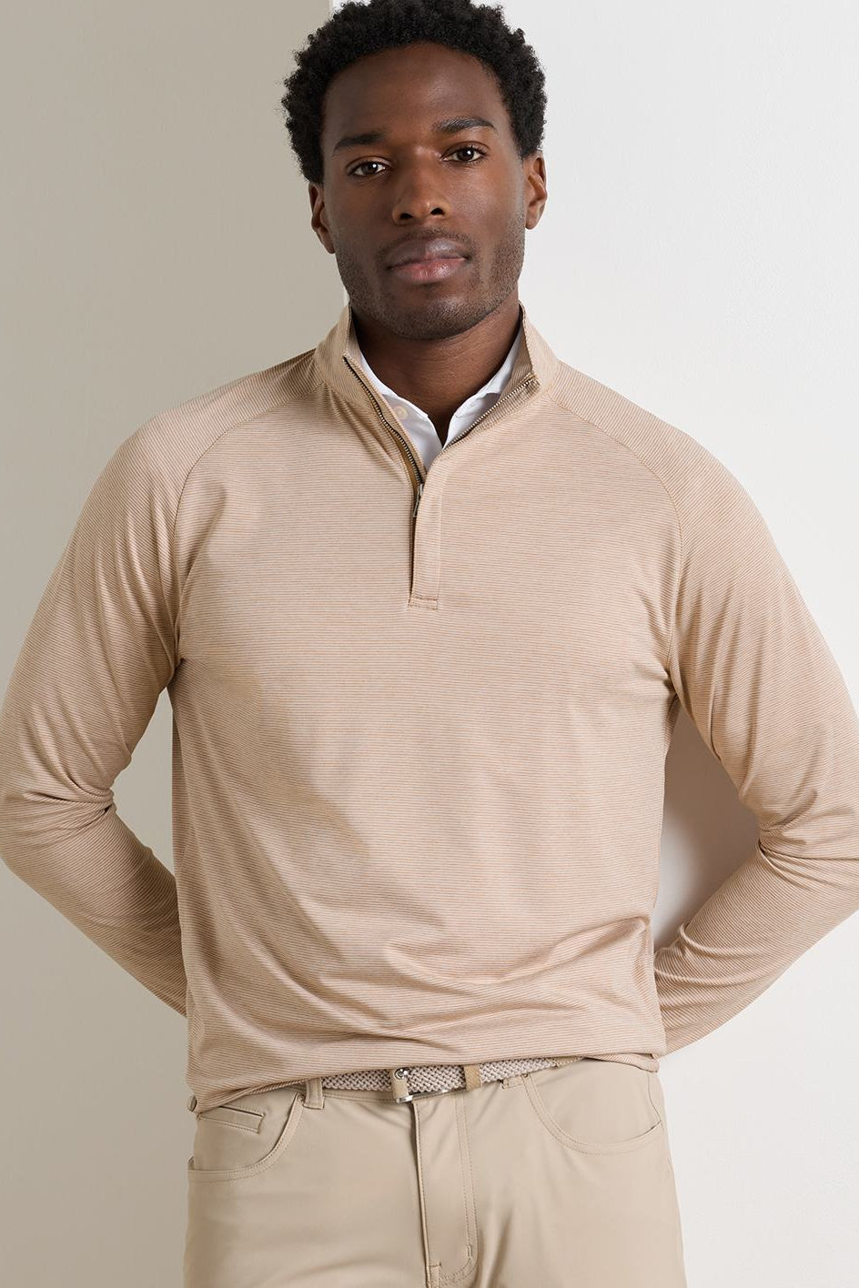 A man wearing a light beige long-sleeve quarter-zip pullover sweater over a white collared shirt, paired with beige trousers, against a plain light gray studio background.