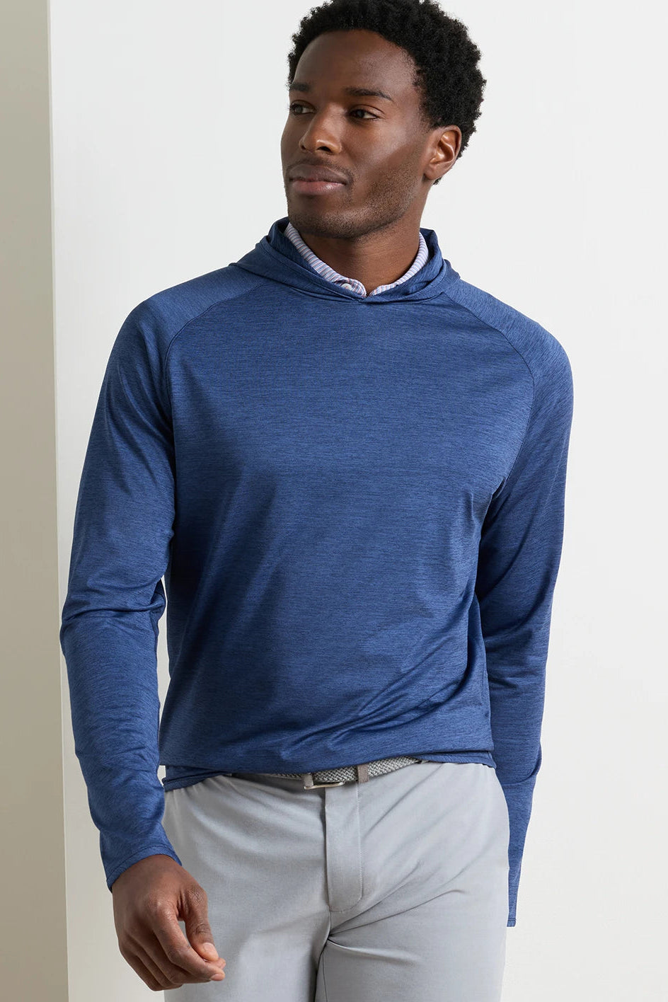 A man wearing a heathered blue long-sleeve quarter-zip pullover hoodie with light gray trousers, standing against a plain off-white wall.