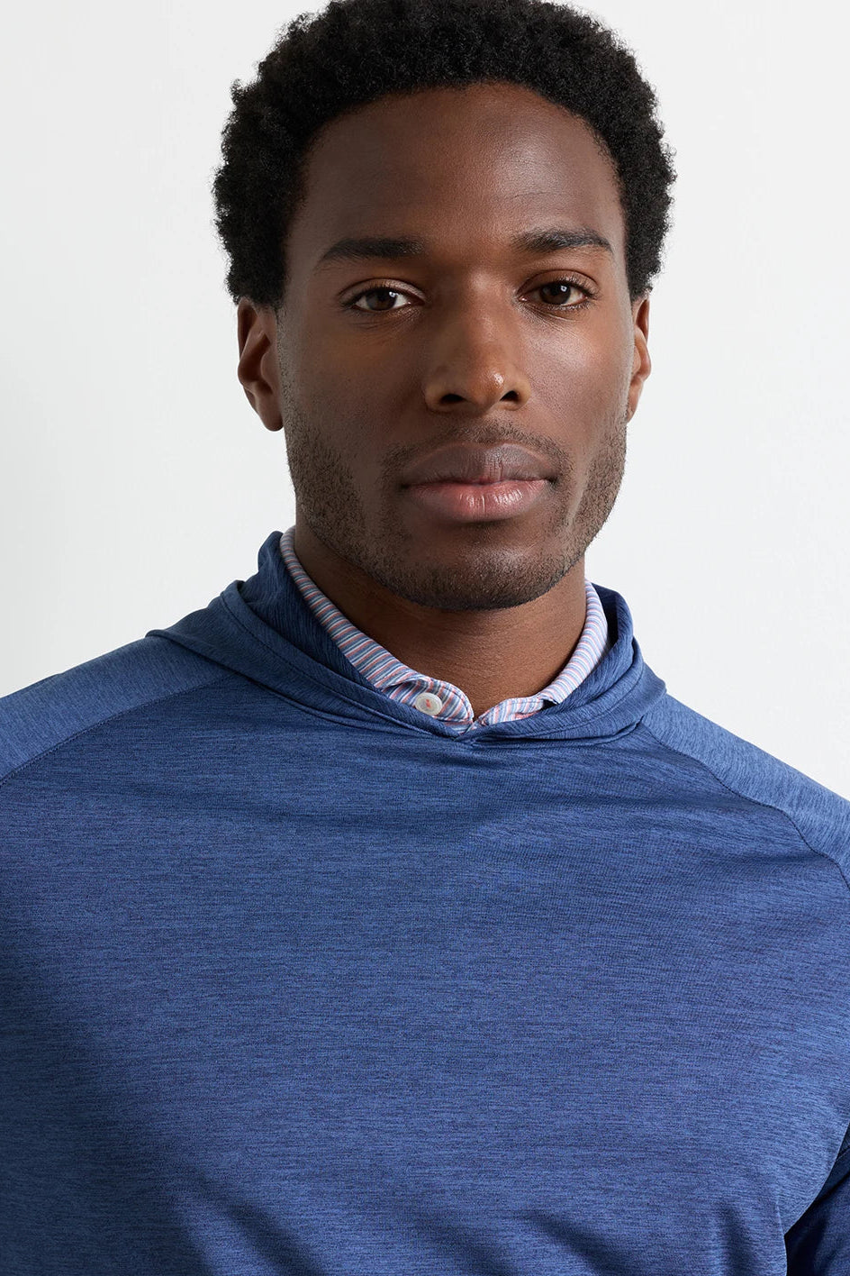 A man wearing a heathered blue athletic pullover hoodie over a striped button-down shirt, shown in a close-up portrait against a light gray background.