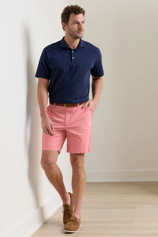 Man wearing a navy polo shirt and pink shorts standing against a plain wall.