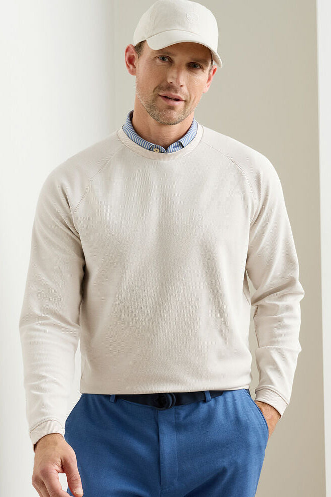 Man wearing a beige sweater and blue pants with a white cap indoors.