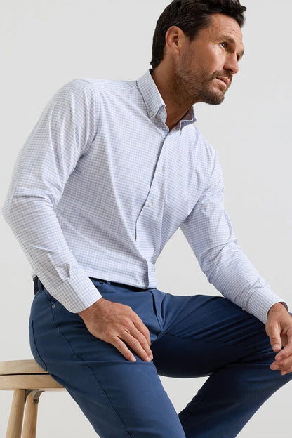 Man wearing a light blue checkered shirt and dark blue pants sitting on a wooden stool against a white background.