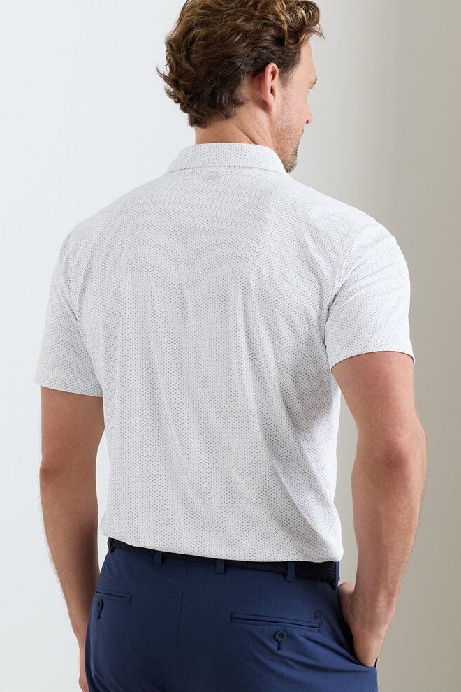 Man wearing a white patterned polo shirt and navy pants against a plain background