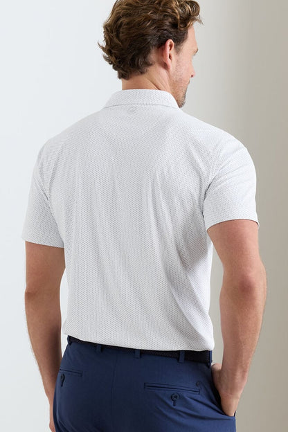 Man wearing a white patterned polo shirt and navy pants against a plain background