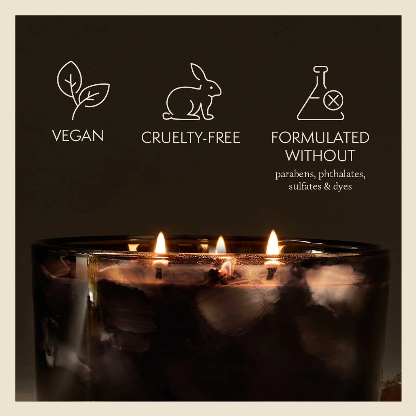 A dark brown triple-wick candle in a clear glass jar with lit flames, set against a dark brown background with white text and icons highlighting 'Vegan', 'Cruelty-Free', and 'Formulated Without' claims.