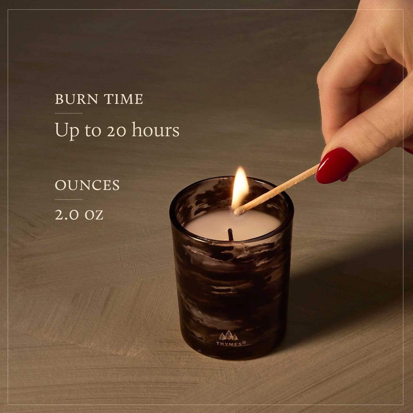 A lit candle in a dark marbled glass jar sits on a wooden surface while a hand with red nail polish holds a match above it. Text on the left reads 'BURN TIME Up to 20 hours' and 'OUNCES 2.