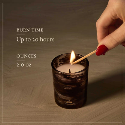 A lit candle in a dark marbled glass jar sits on a wooden surface while a hand with red nail polish holds a match above it. Text on the left reads 'BURN TIME Up to 20 hours' and 'OUNCES 2.