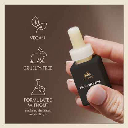 Hand holding a Thymes Noir Woods essential oil dropper with vegan, cruelty-free, and sulfate-free icons on a brown background.