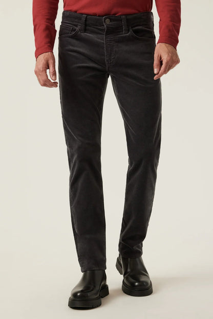 A person wearing slim-fit black jeans, a red long-sleeve top, and black leather boots, standing against a plain light studio background.