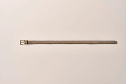 A long, slim leather belt in a muted taupe-brown color with a silver-toned metal buckle, laid horizontally on a plain off-white background.