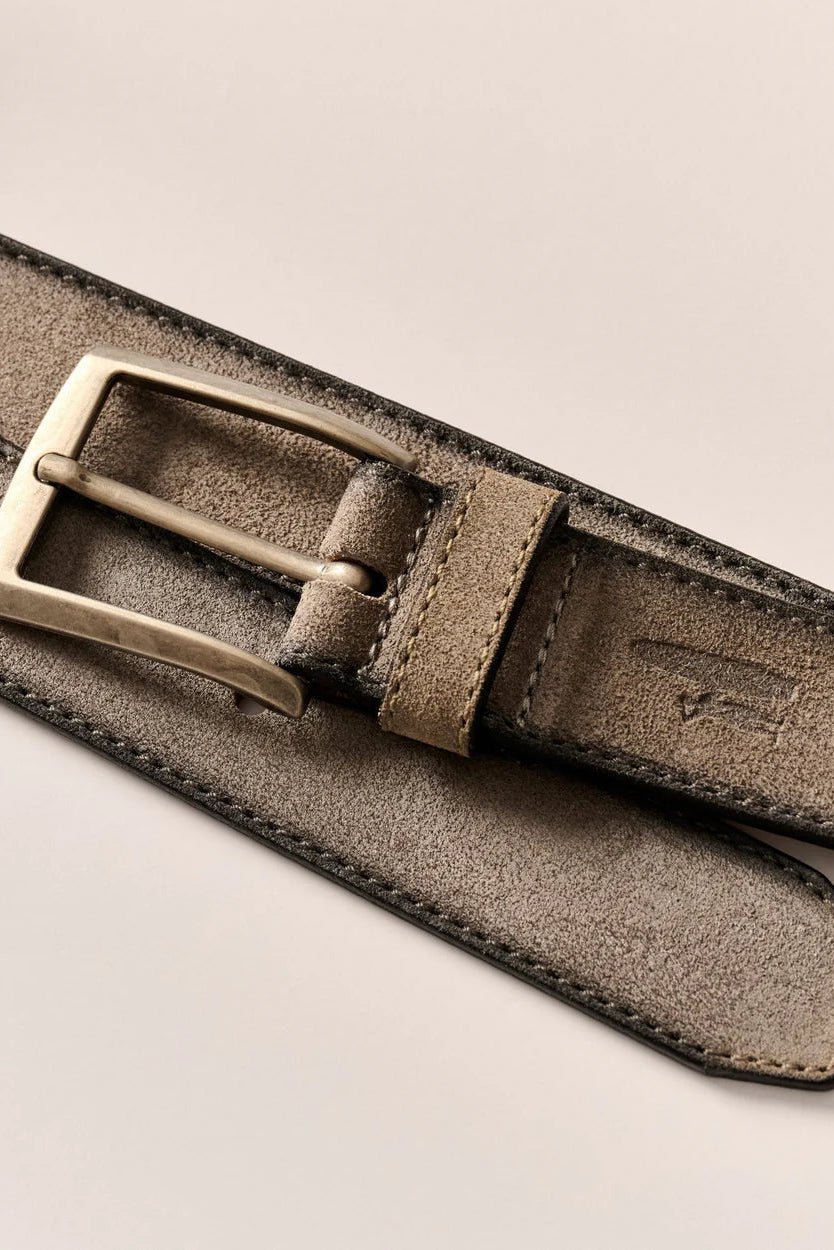 A close-up of a taupe suede belt with a brushed brass buckle, laid diagonally on a light beige background.