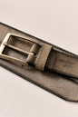 A close-up of a taupe suede belt with a brushed brass buckle, laid diagonally on a light beige background.