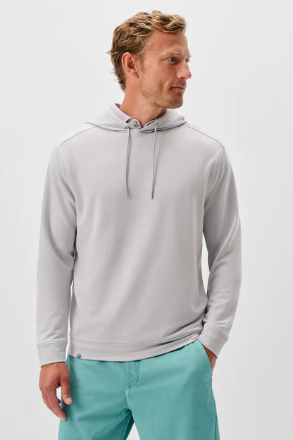 A man wearing a light gray pullover hoodie with matching gray drawstrings, paired with teal pants, standing against a light gray studio background.