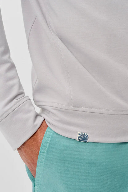 Close-up of a person wearing a light gray long-sleeve top and teal shorts, with a small blue and white logo patch near the hem of the top.