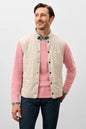 Man wearing a pink sweater with a beige quilted vest on a light gray background