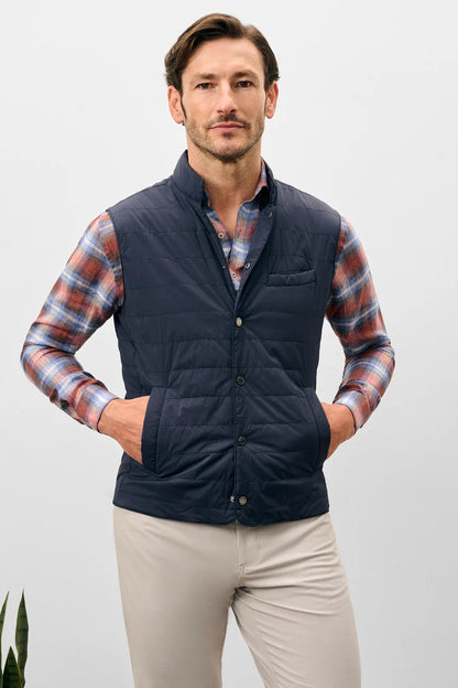 Man wearing a navy puffer vest over a plaid shirt with beige pants on a light gray background