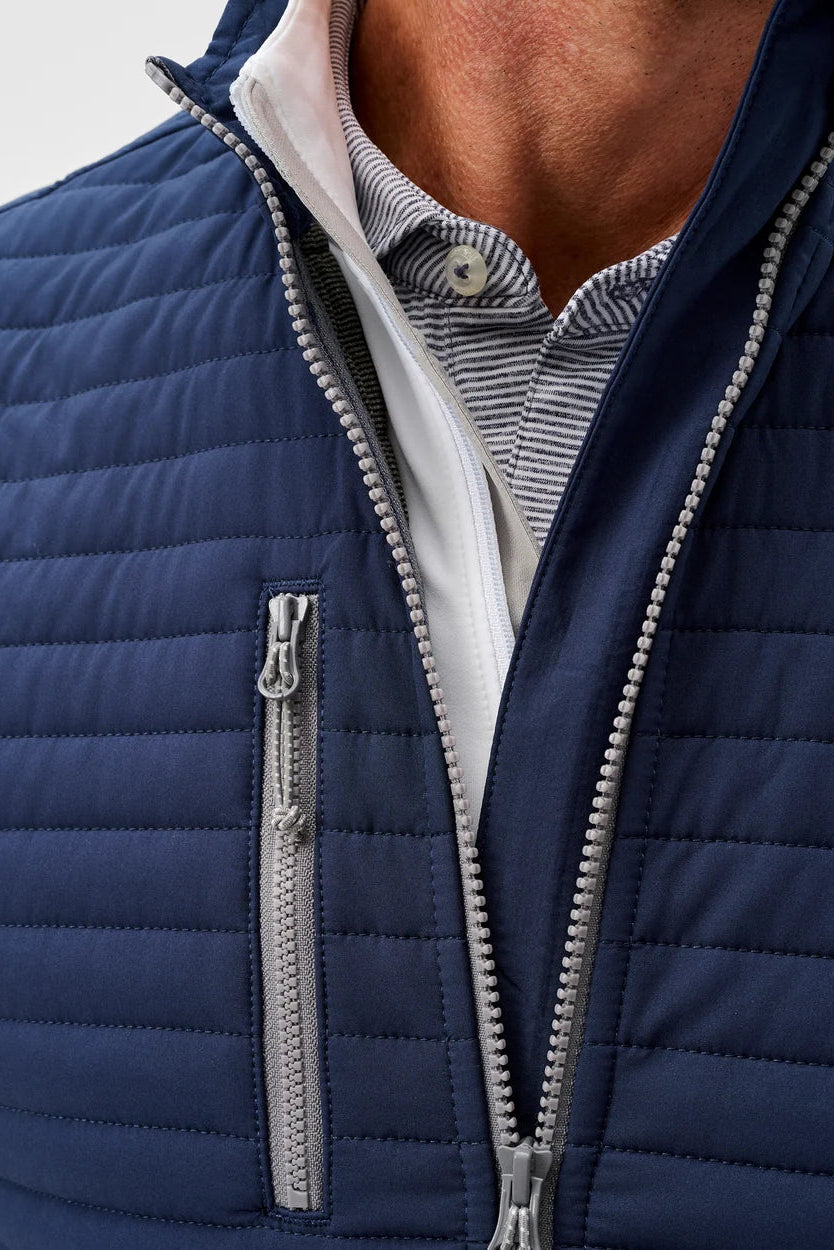 Close-up of a navy blue quilted zip-front jacket worn over a striped collared shirt, showing the zipper, chest pocket, and part of the model's neck and chest.