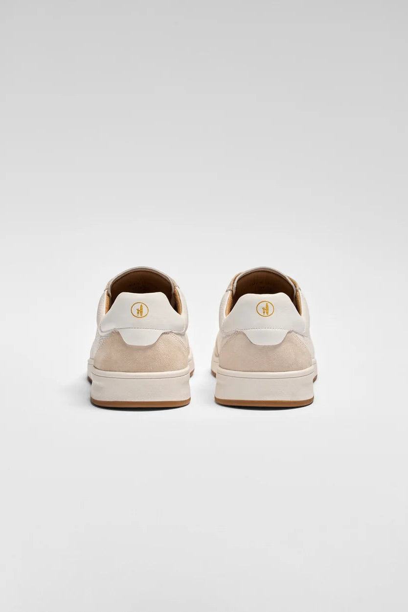 A pair of low-top sneakers shown from the back, featuring cream suede and white leather panels with a small gold circular logo on the heel, set against a light gray studio background.
