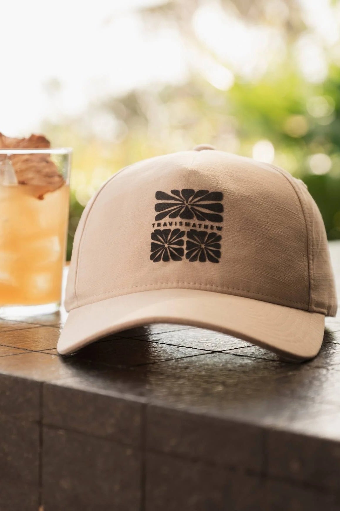 Beige cap with a black logo on a wooden surface next to a glass of iced tea.