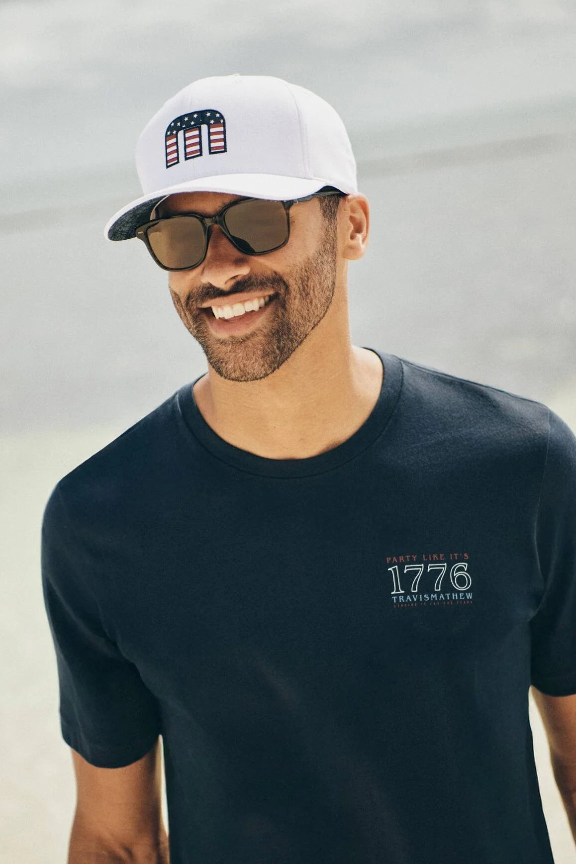Man wearing a black t-shirt with '1776' logo, white cap, and sunglasses on a beach.