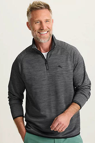 A smiling man wearing a dark gray heathered quarter-zip pullover sweater with a small embroidered logo on the left chest, paired with green pants, against a plain light background.