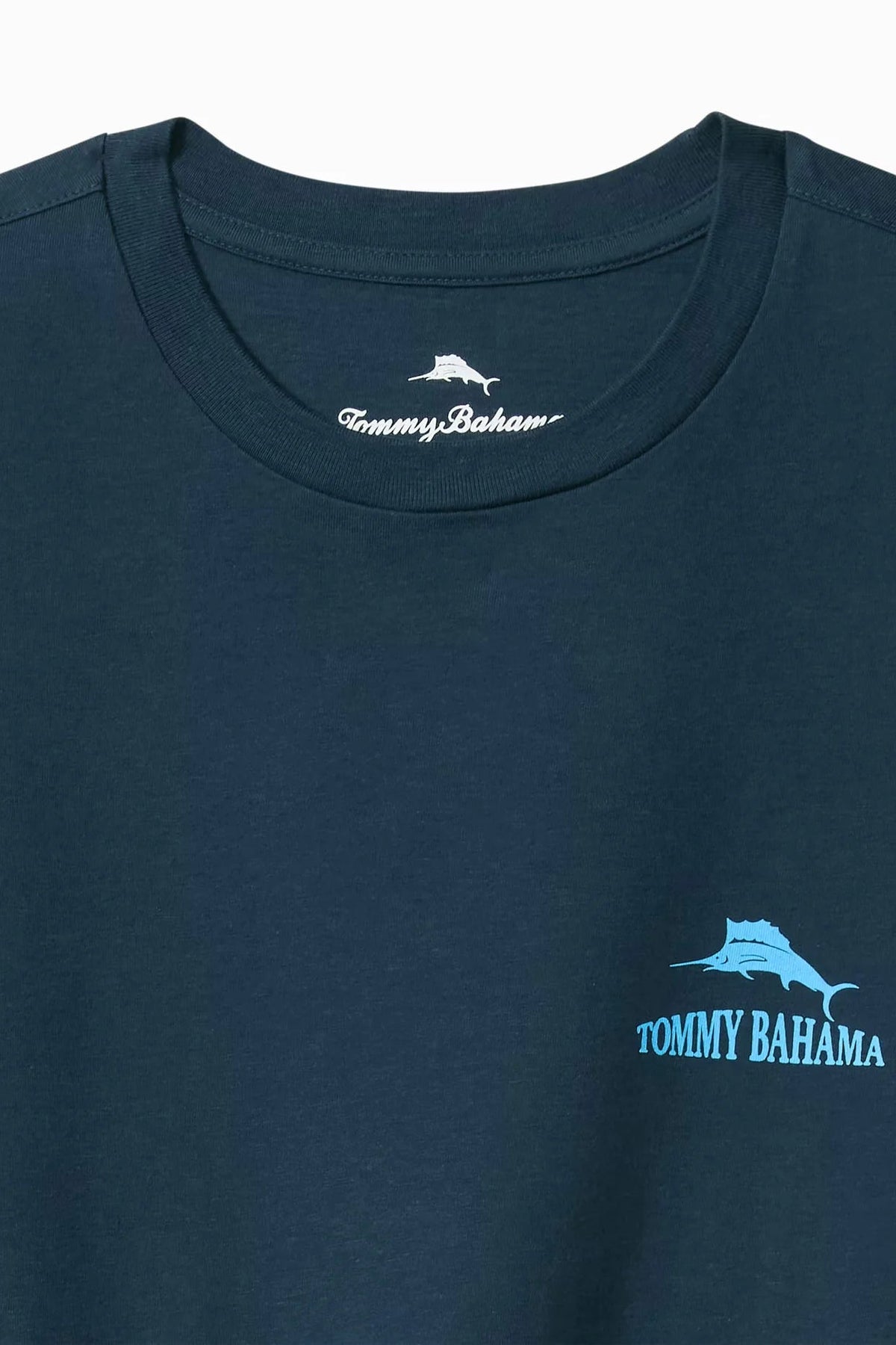 Close-up of a dark navy blue Tommy Bahama t-shirt featuring a light blue marlin graphic and brand name on the left chest, with the white Tommy Bahama neck label visible inside the collar.