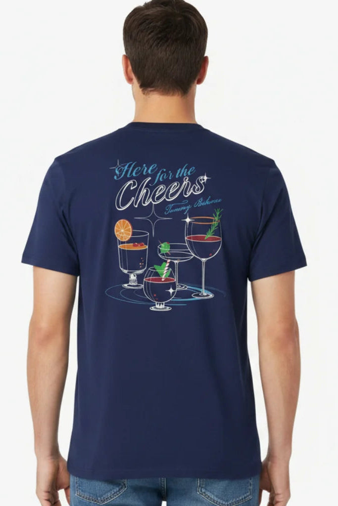 A man viewed from the back wearing a navy blue t-shirt with a colorful graphic of cocktail glasses and the text 'Here for the Cheers' on the back, paired with blue jeans against a plain white background.