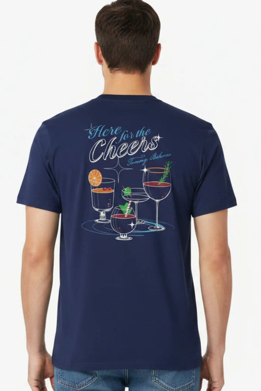 A man viewed from the back wearing a navy blue t-shirt with a colorful graphic of cocktail glasses and the text 'Here for the Cheers' on the back, paired with blue jeans against a plain white background.