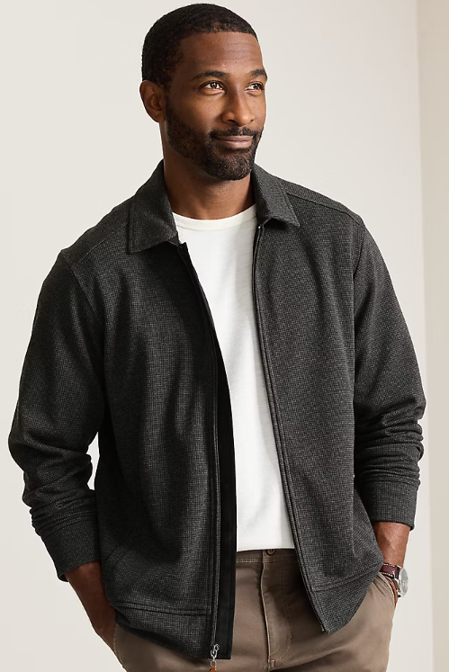 A man wearing a dark charcoal textured zip-up jacket over a white t-shirt and khaki pants, standing against a neutral light beige background.