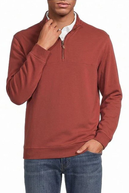A man wearing a rust-colored quarter-zip pullover sweater over a white collared shirt and blue jeans, shown from the chin down against a plain white background.