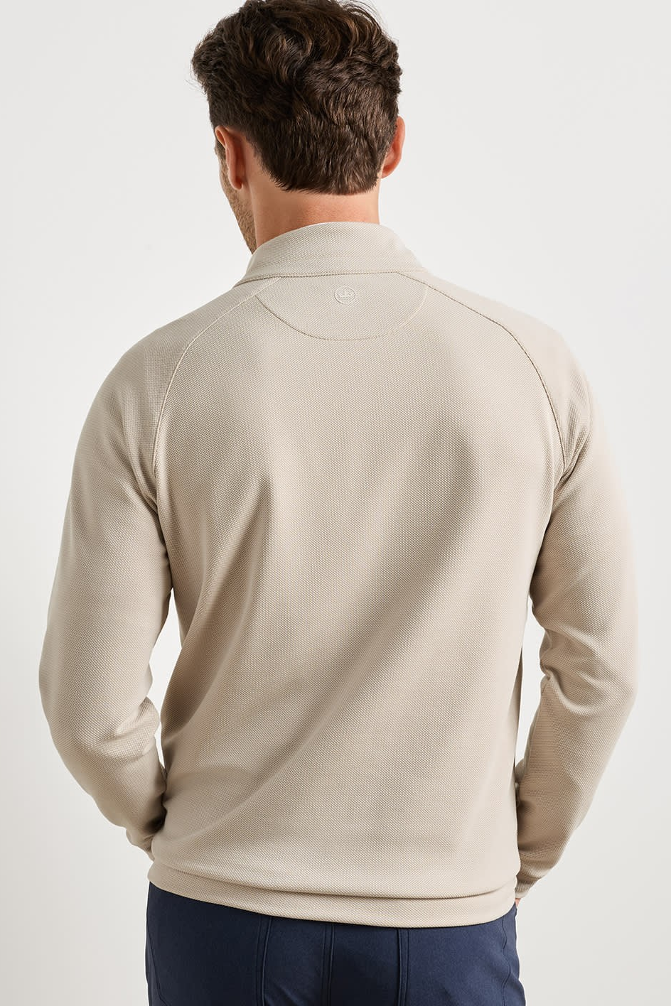 A man viewed from behind wearing a light beige long-sleeve quarter-zip pullover sweatshirt and navy blue pants, set against a plain light gray studio background.