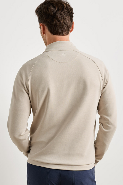 A man viewed from behind wearing a light beige long-sleeve quarter-zip pullover sweatshirt and navy blue pants, set against a plain light gray studio background.