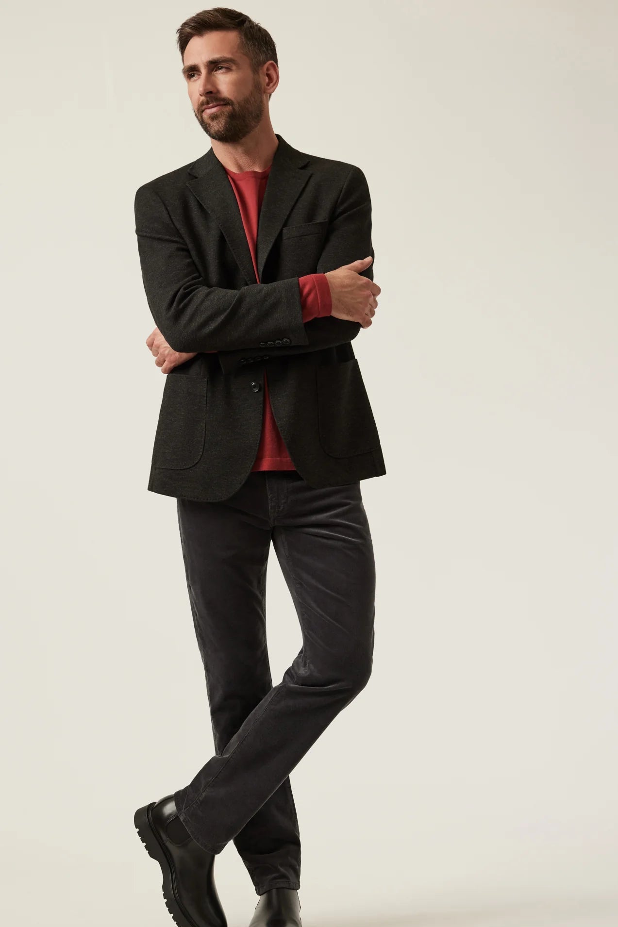 A man modeling a black blazer over a red shirt, paired with dark charcoal trousers and black shoes, standing against a plain light beige studio background.