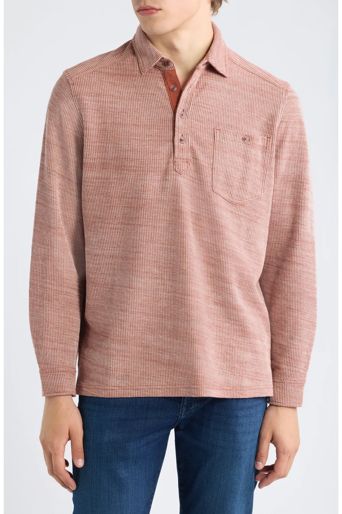 A man wearing a dusty rose long-sleeve polo shirt with a textured weave and a chest pocket, paired with blue jeans, against a light gray background.
