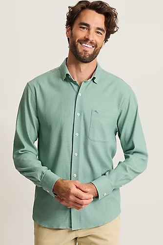 A smiling man wearing a sage green long-sleeve button-down shirt with a chest pocket, paired with khaki pants, against a plain light beige studio background.