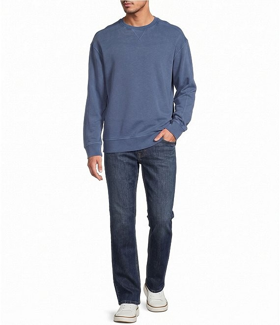 A man wearing a dusty blue crewneck sweatshirt, dark blue jeans, and white sneakers, standing against a plain white background.