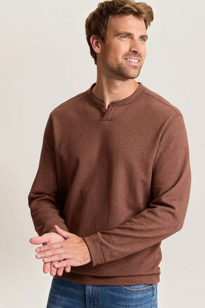 A man wearing a brown long-sleeve henley sweatshirt and blue jeans, standing against a light gray studio background.