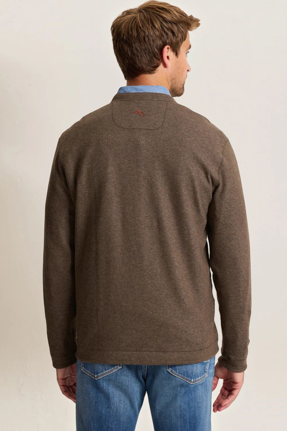 A man viewed from the back wearing a brown knit quarter-zip sweater over a light blue collared shirt and blue jeans, against a plain off-white studio background.