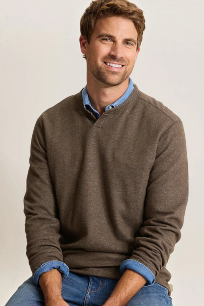 A smiling man wearing a brown long-sleeve knit sweater over a light blue collared shirt and blue jeans, seated against a plain light gray background.