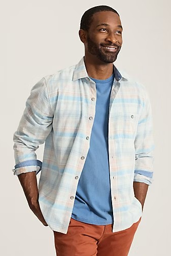 A man wearing a light blue and white plaid button-down shirt over a blue t-shirt and rust-orange pants, photographed against a solid light beige background.
