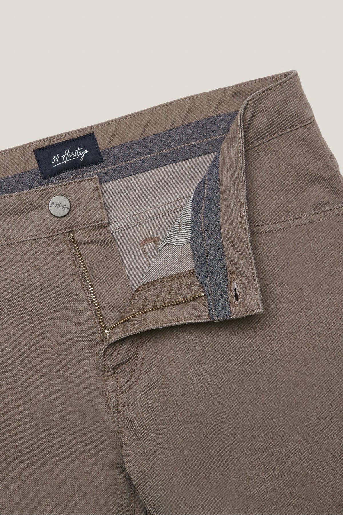 Close-up of a pair of St. Hilda men's trousers in a taupe-brown color, showing the interior waistband with a patterned lining, a silver metal button, and a zippered pocket.