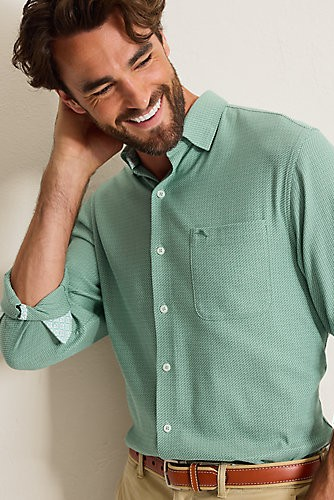 A man wearing a light green textured long-sleeve button-down shirt with rolled-up sleeves and khaki pants, posing against a neutral beige wall.