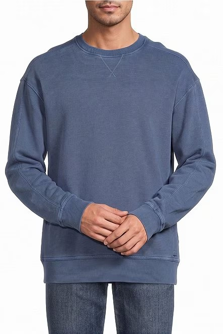 A man wearing a muted slate blue crewneck sweatshirt and dark blue jeans, standing against a plain white background.