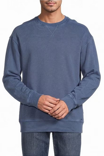 A man wearing a muted slate blue crewneck sweatshirt and dark blue jeans, standing against a plain white background.