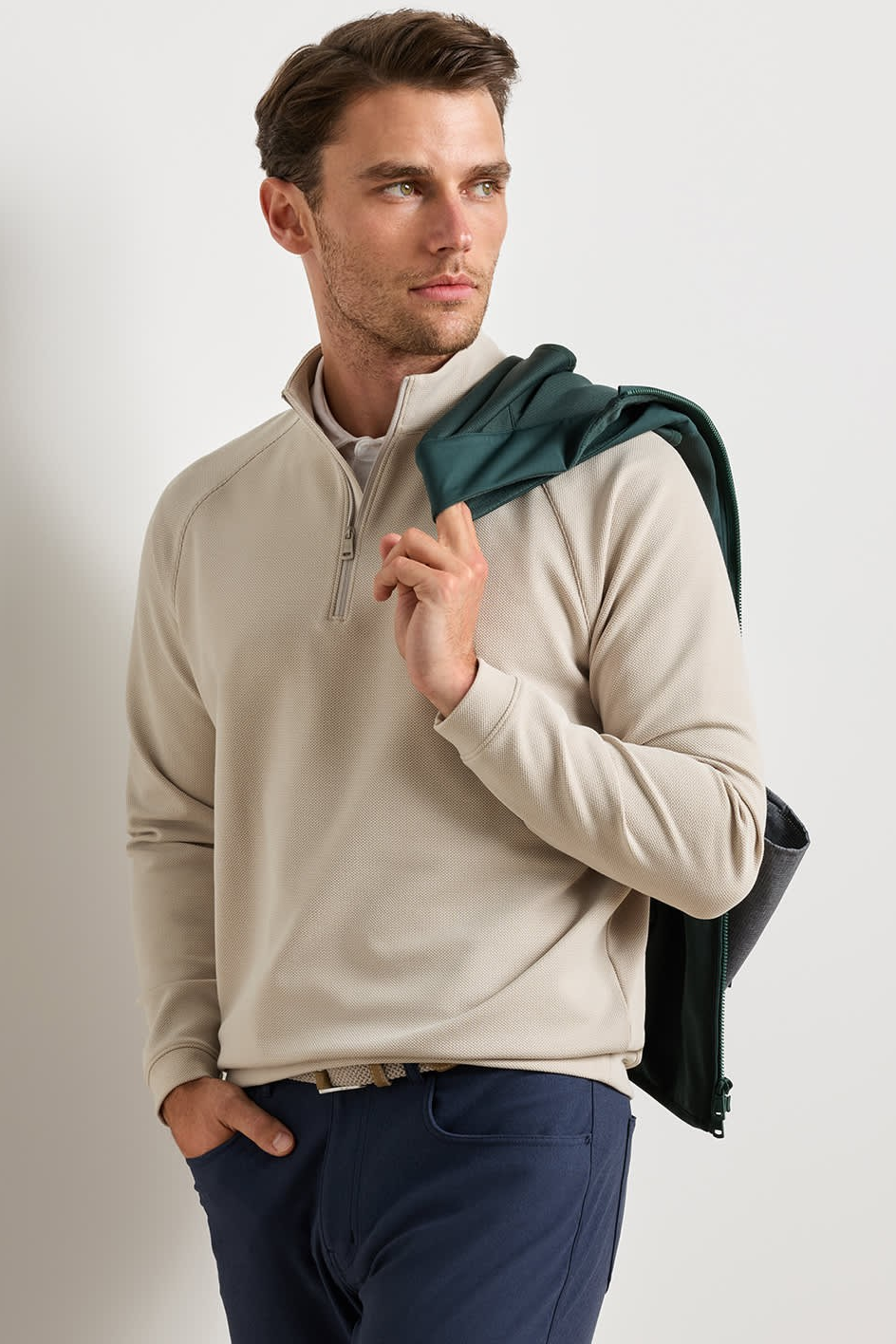 A man wearing a light beige quarter-zip pullover sweater and navy trousers, with a dark green jacket draped over his shoulder, set against a plain light gray studio background.
