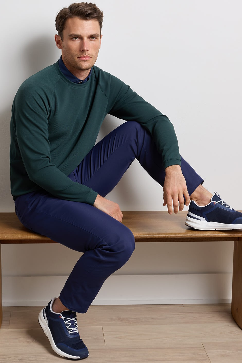 A man sits on a wooden bench wearing a dark green mock-neck long-sleeve shirt, navy blue trousers, and navy blue athletic sneakers against a light gray wall.